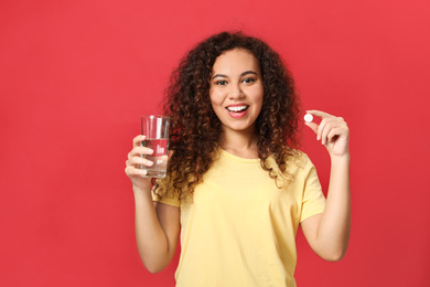 African-American woman with glass of water and vitamin pill on red background