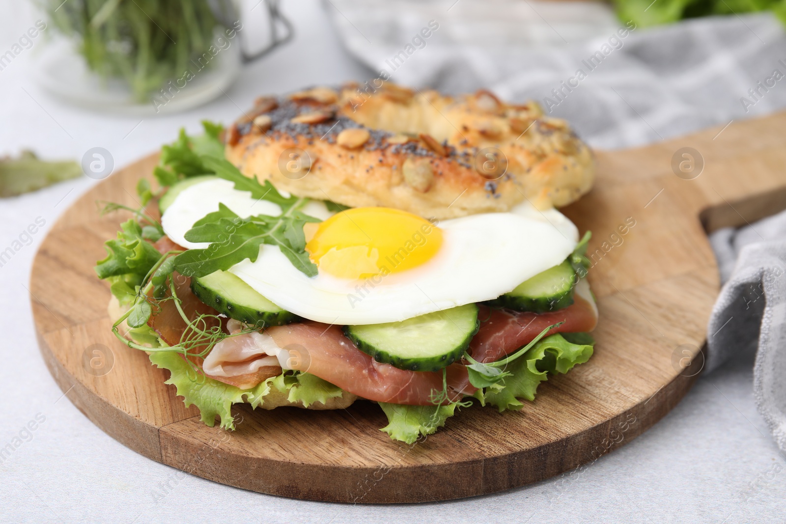 Photo of Tasty bagel with cured ham, egg, cucumber and salad mix on light table, closeup