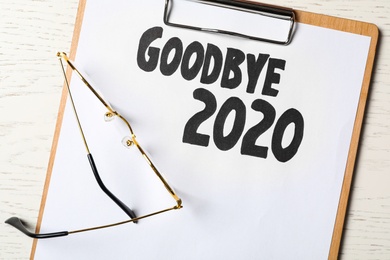 Clipboard with phrase Goodbye 2020 and glasses on white wooden table, flat lay