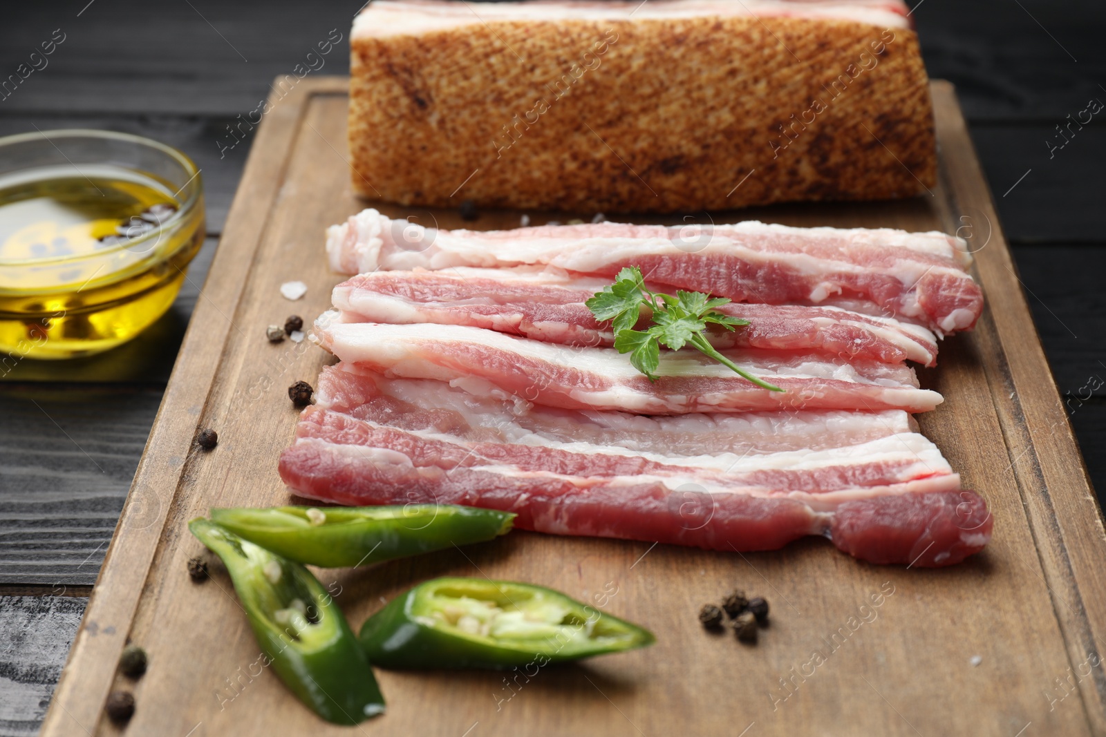 Photo of Pieces of raw pork belly, chili pepper, peppercorns, rosemary and parsley on black wooden table, closeup