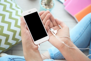Photo of Young woman holding mobile phone with blank screen and earphones in hands, indoors