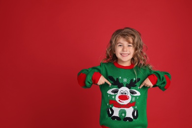 Photo of Cute little girl pointing at her green Christmas sweater against red background. Space for text