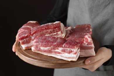 Photo of Woman holding wooden board with pieces of raw pork belly on black background, closeup