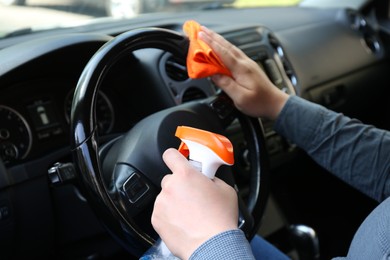 Photo of Man cleaning steering wheel with rag and spray bottle in car, closeup