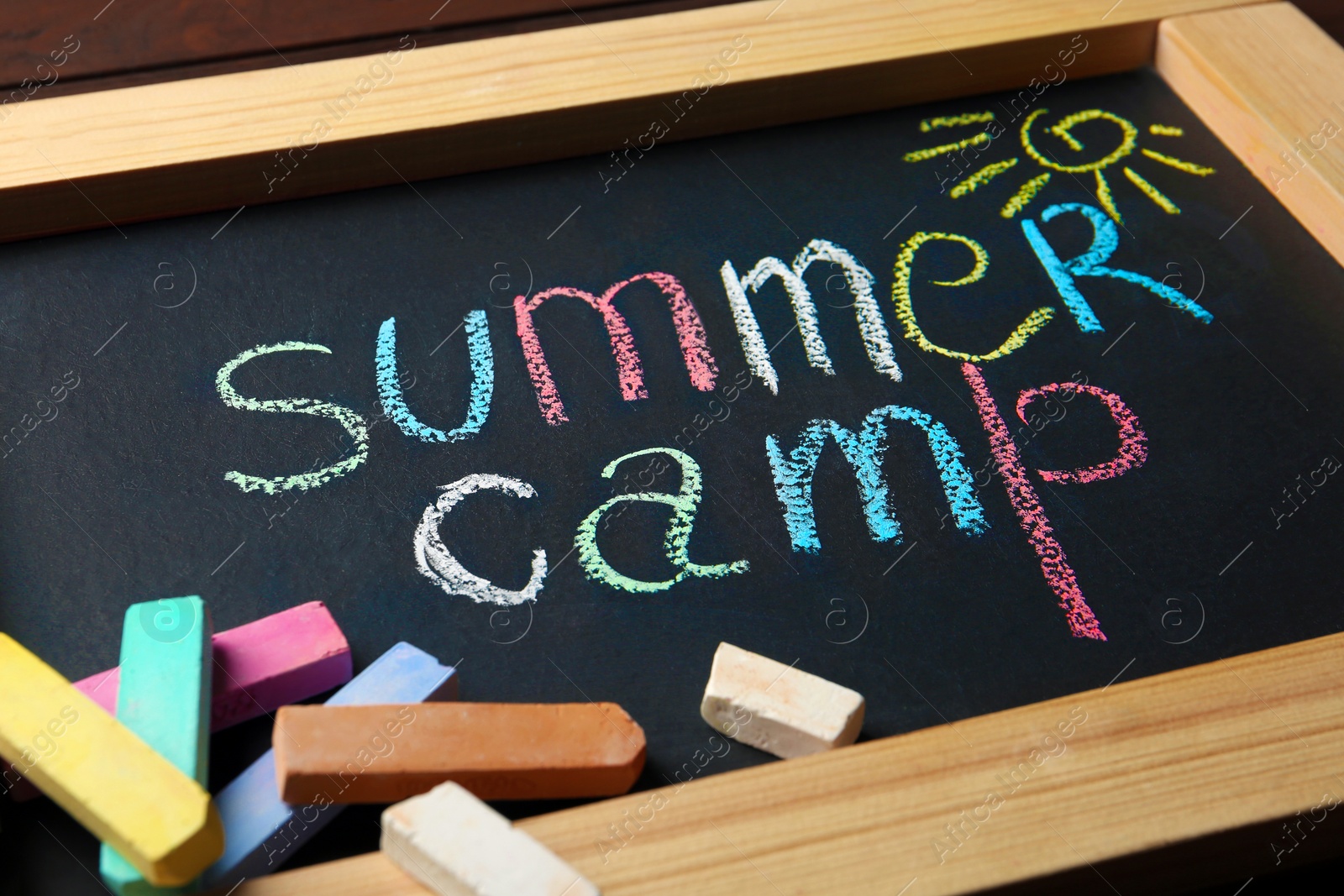 Photo of Small blackboard with text SUMMER CAMP, drawing and chalk sticks on wooden background, closeup