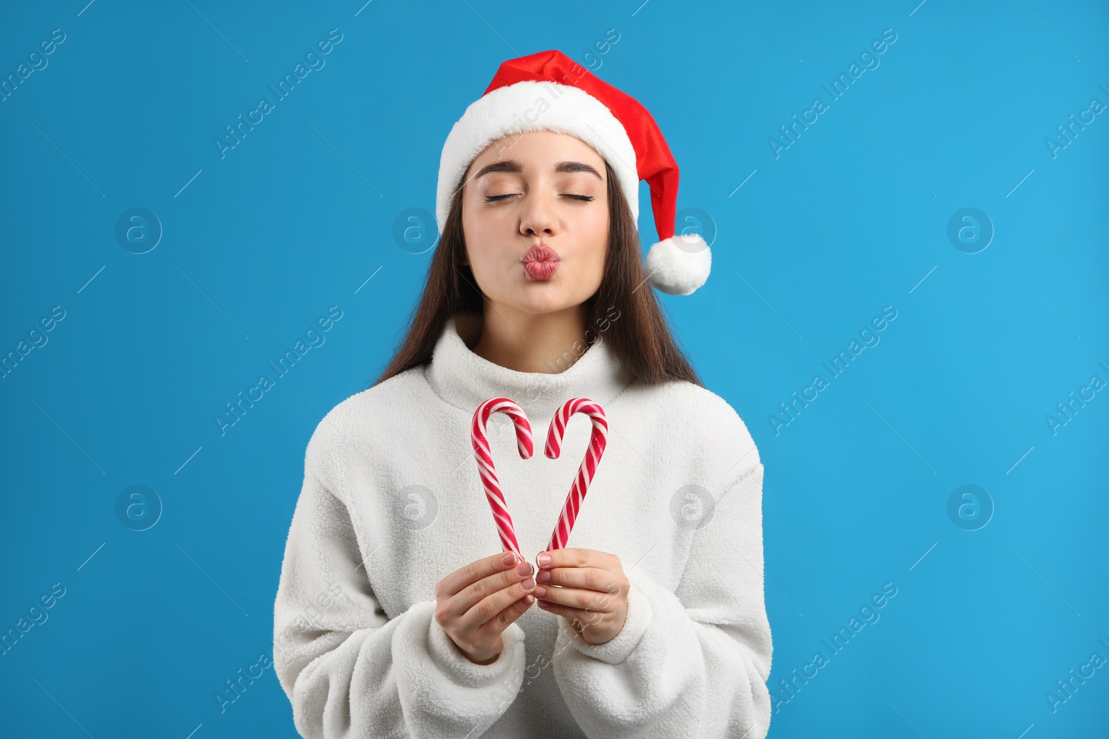 Photo of Young woman in beige sweater and Santa hat holding candy canes on blue background. Celebrating Christmas