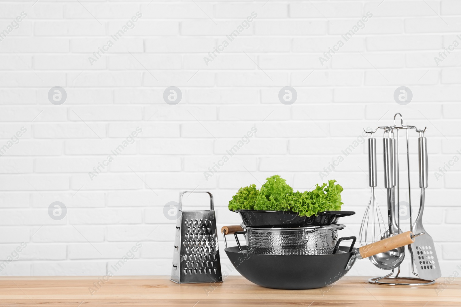 Photo of Set of clean cookware, utensils and lettuce on table against  white brick wall. Space for text
