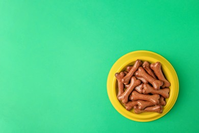 Photo of Yellow bowl with bone shaped dog cookies on green background, top view. Space for text