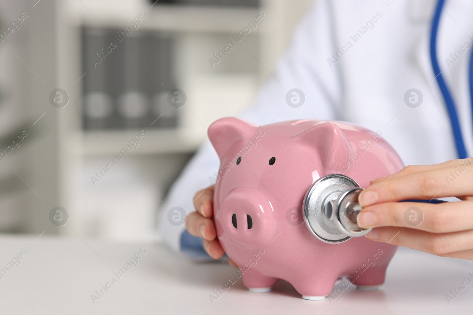 Photo of Doctor with piggy bank at white table indoors, closeup. Space for text