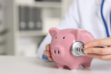 Doctor with piggy bank at white table indoors, closeup. Space for text