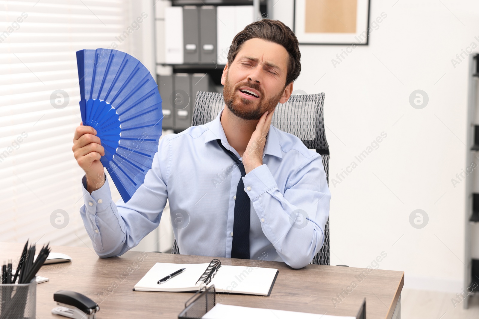 Photo of Bearded businessman waving blue hand fan to cool himself at table in office
