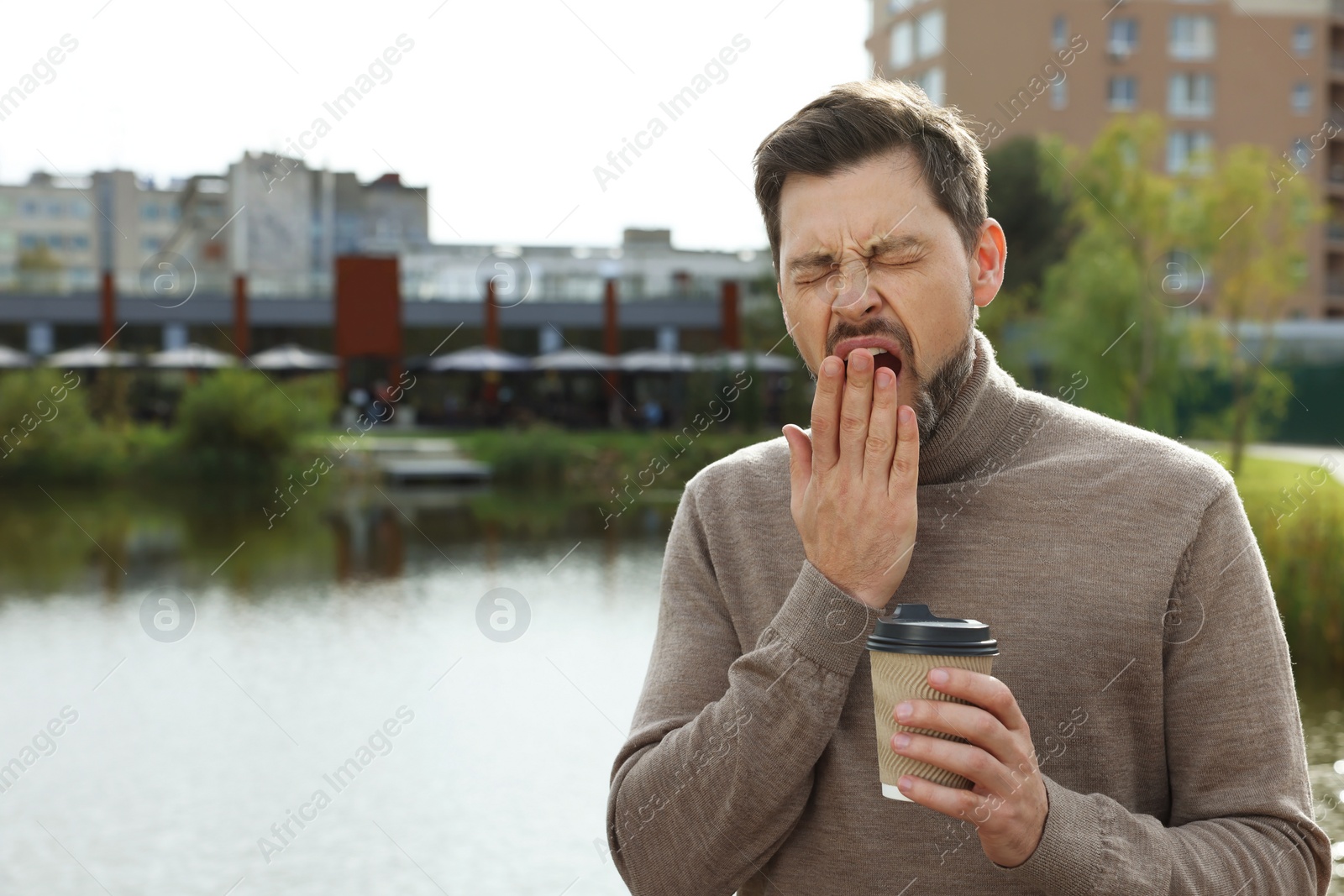 Photo of Sleepy man with cup of coffee yawning near river outdoors. Space for text