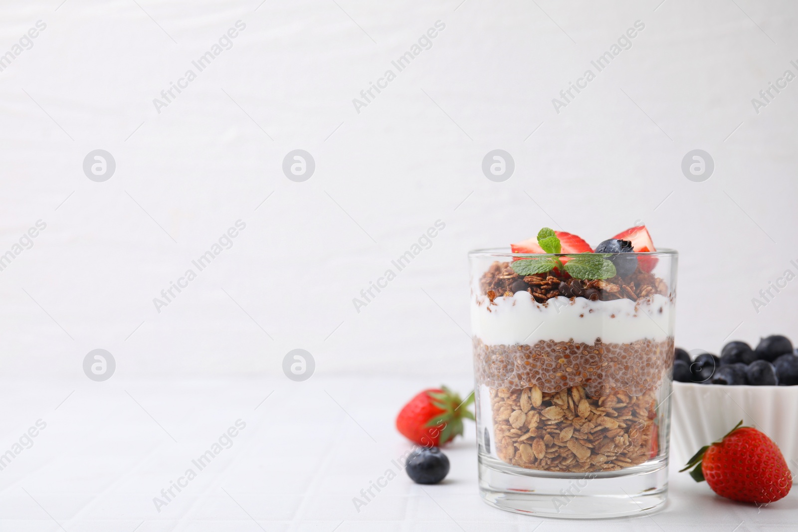 Photo of Tasty granola with berries, yogurt and chia seeds in glass on white tiled table, space for text