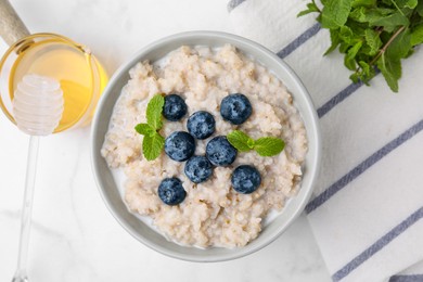 Photo of Delicious barley porridge with blueberries and mint served with honey on white marble table, flat lay
