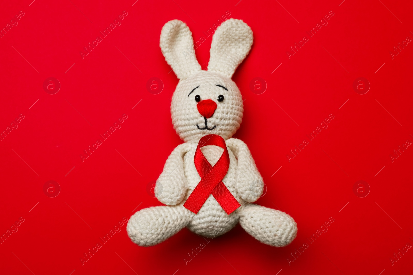 Photo of Cute knitted toy bunny with ribbon on red background, top view. AIDS disease awareness