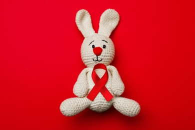 Cute knitted toy bunny with ribbon on red background, top view. AIDS disease awareness