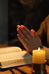 Photo of Woman praying at table with burning candle and Bible, closeup