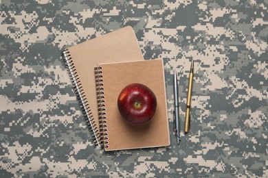 Photo of Notebooks, pens and apple on camouflage background, flat lay. Military education