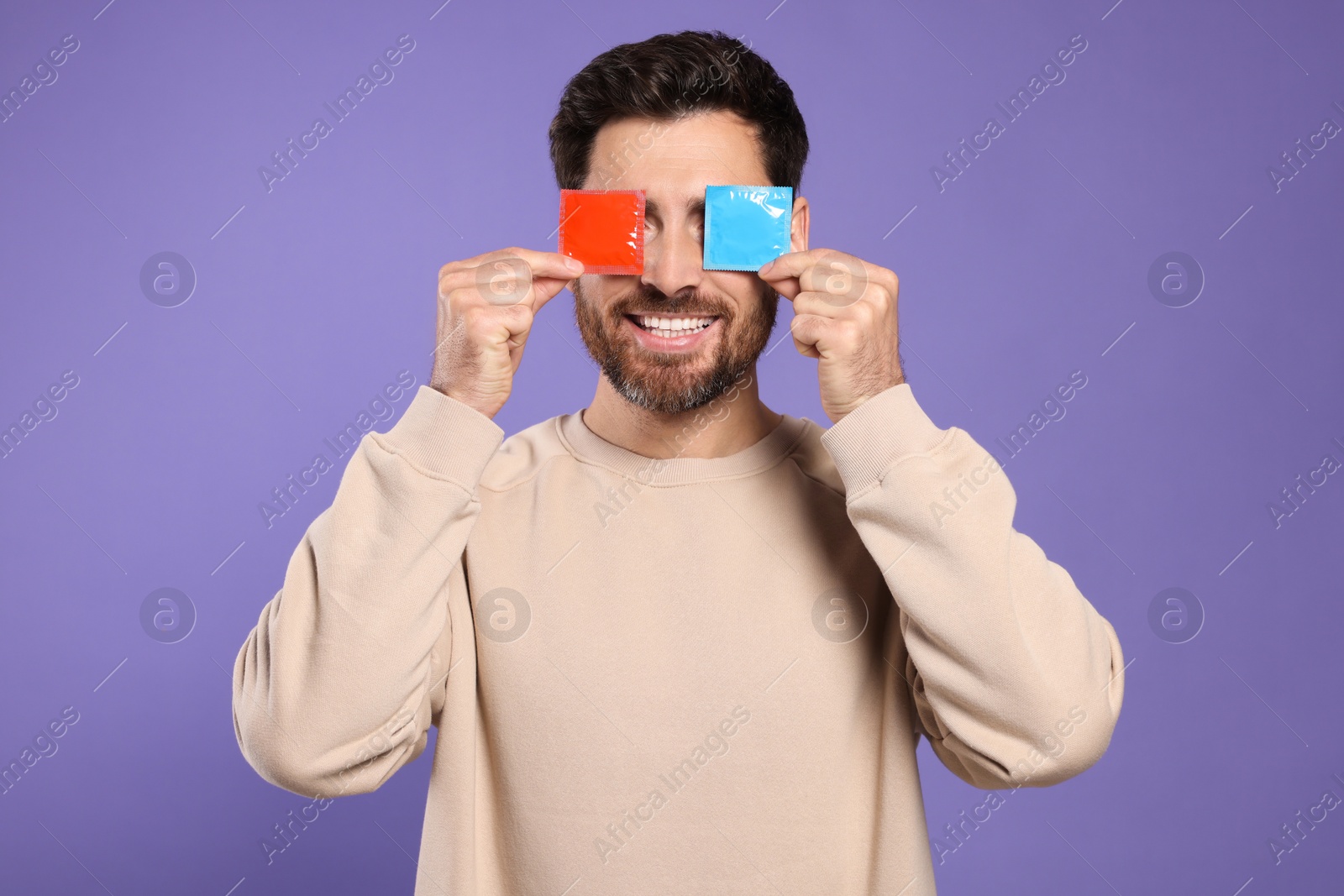 Photo of Man holding condoms near his eyes on purple background. Safe sex