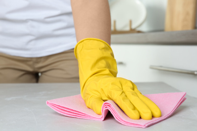 Photo of Woman in gloves wiping grey table with rag indoors, closeup