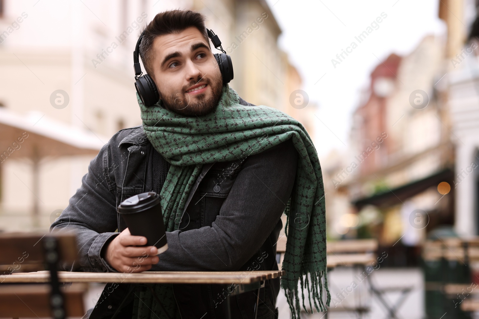 Photo of Smiling man in warm scarf with paper cup listening to music in outdoor cafe