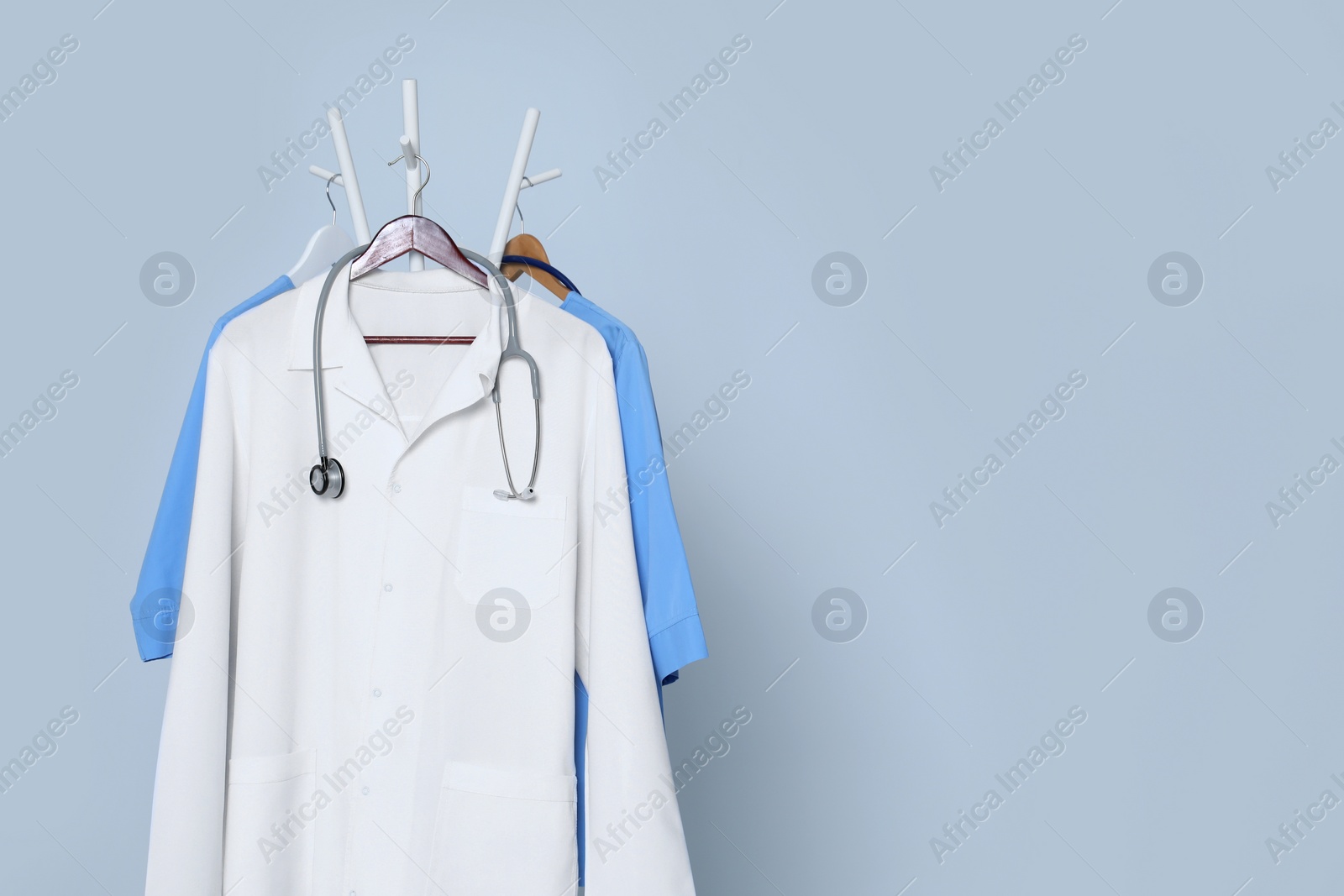 Photo of Medical uniforms and stethoscope hanging on rack against light grey background. Space for text