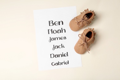 Photo of List of baby names and child's shoes on beige background, flat lay