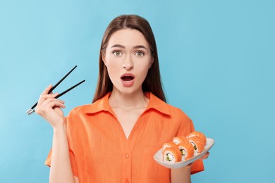 Emotional young woman with plate of sushi rolls and chopsticks on light blue background