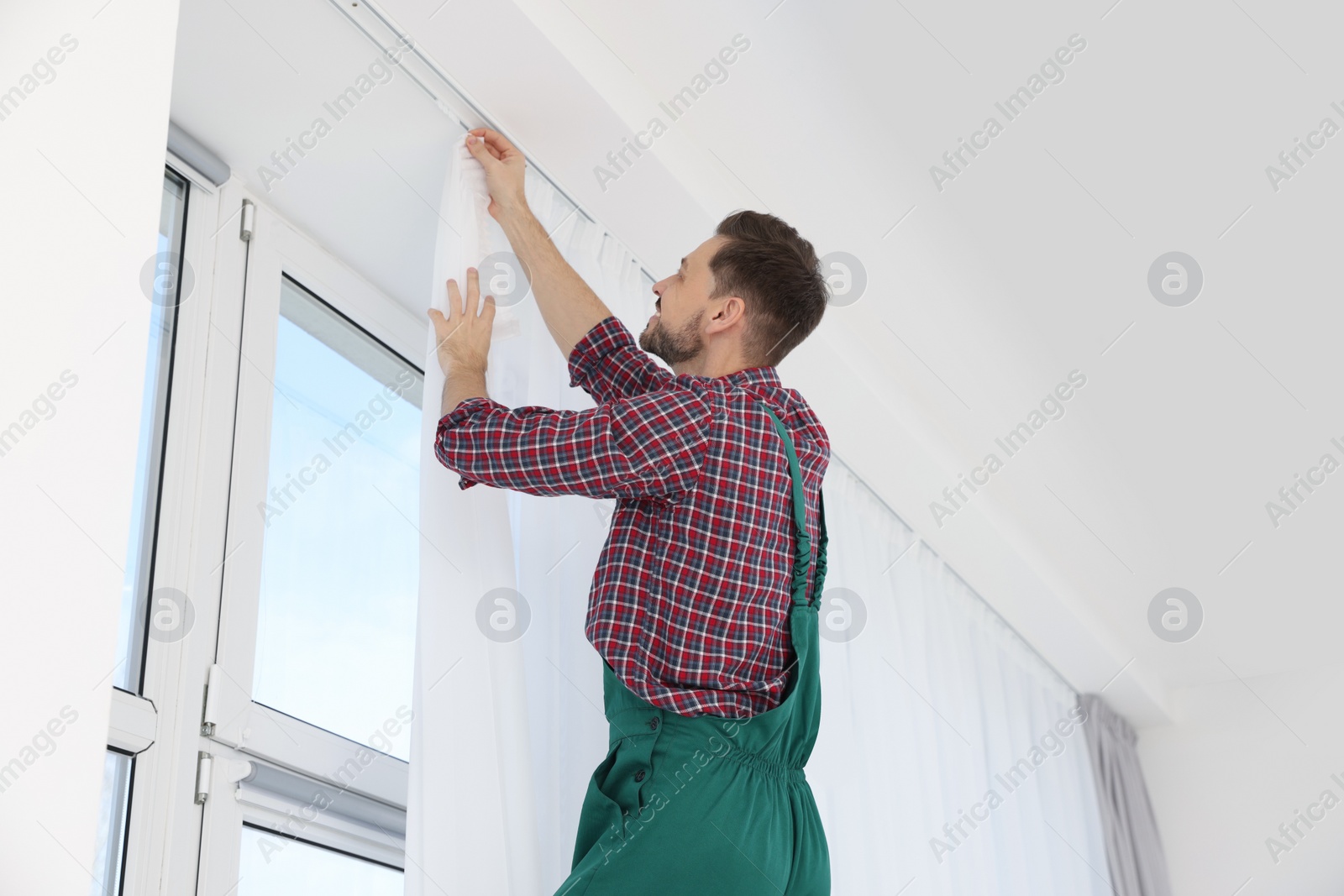 Photo of Worker in uniform hanging window curtain indoors, low angle view