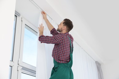 Photo of Worker in uniform hanging window curtain indoors, low angle view