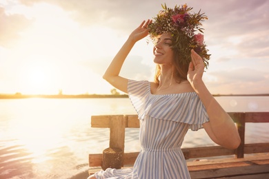 Photo of Young woman wearing wreath made of beautiful flowers on pier near river