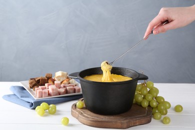 Photo of Woman dipping piece of bread into fondue pot with tasty melted cheese at white wooden table against gray background, closeup