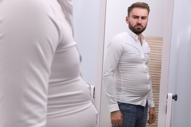 Photo of Upset man wearing tight shirt in front of mirror at home. Overweight problem