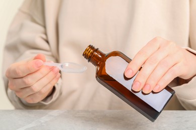 Photo of Woman pouring syrup from bottle into dosing spoon at table, closeup. Cold medicine
