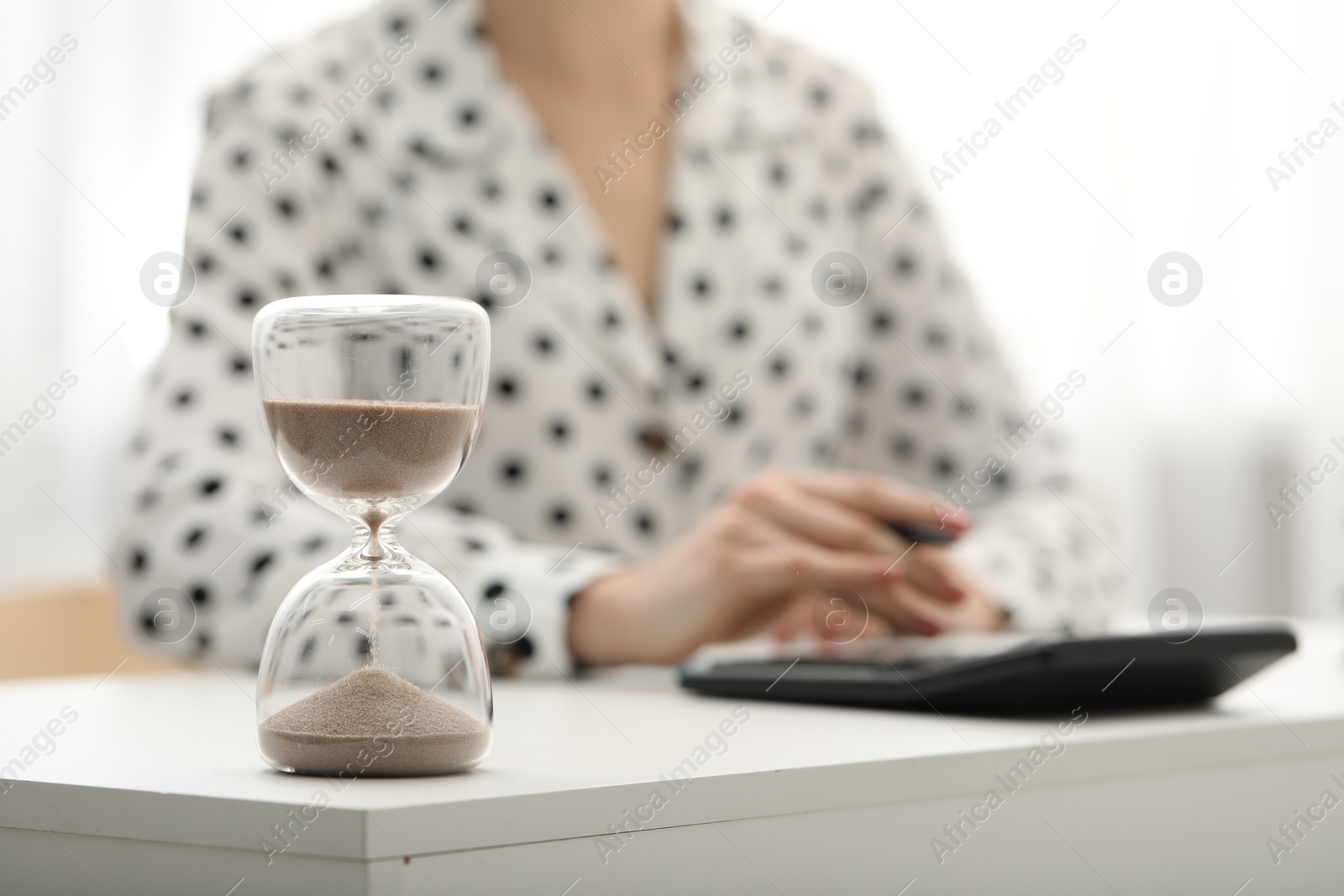 Photo of Hourglass with flowing sand on desk. Woman taking notes while using calculator indoors, selective focus