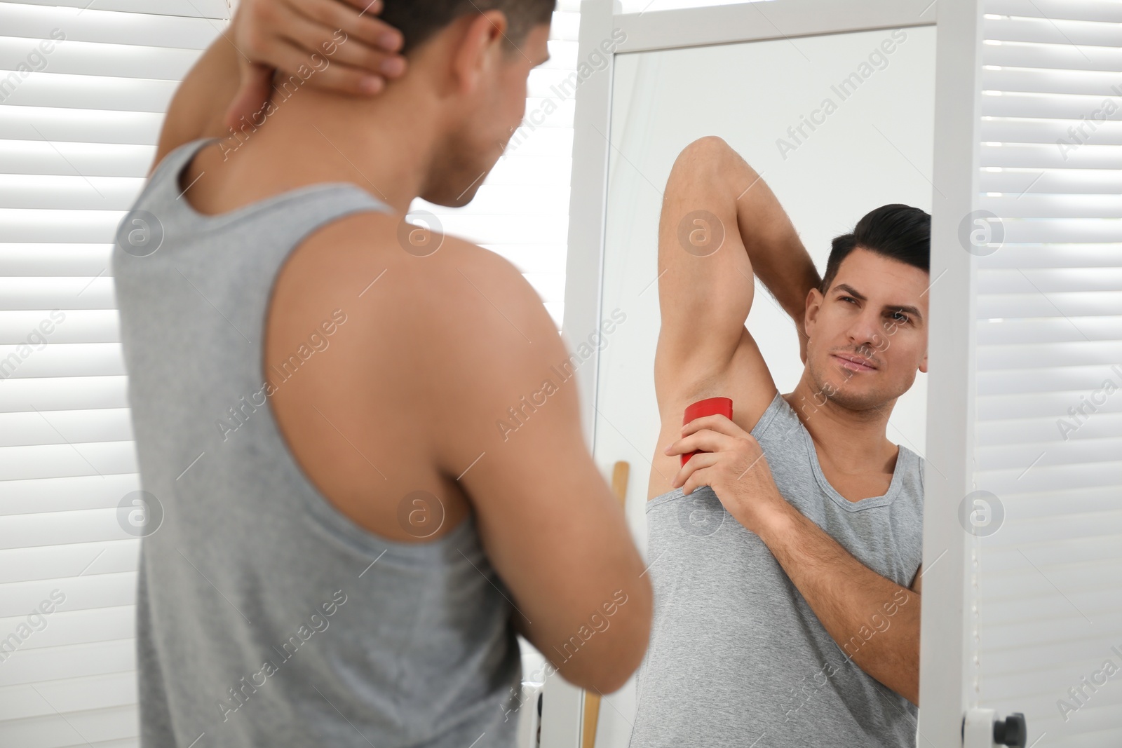 Photo of Handsome man applying deodorant to armpit near mirror at home