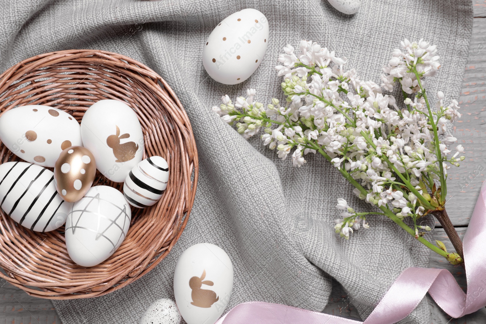 Photo of Many painted Easter eggs, branch with lilac flowers and ribbon on grey wooden table, flat lay