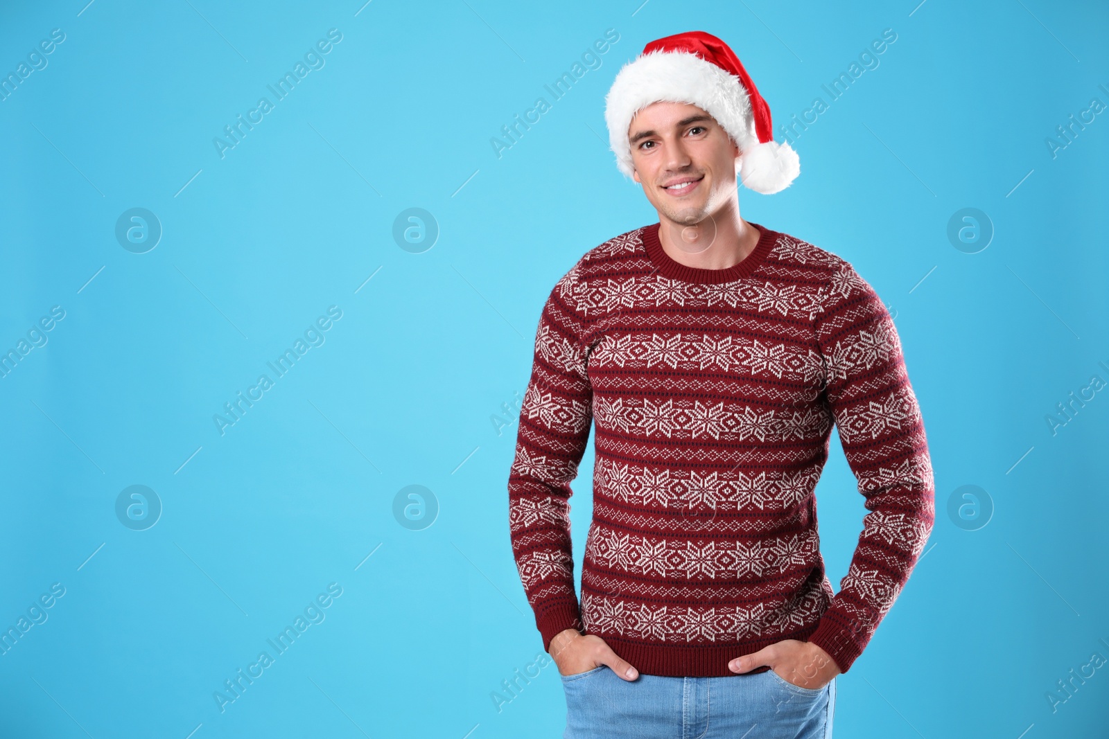 Photo of Portrait of young man in Christmas sweater and Santa hat on light blue background. Space for text