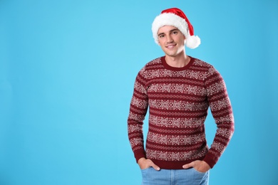 Portrait of young man in Christmas sweater and Santa hat on light blue background. Space for text