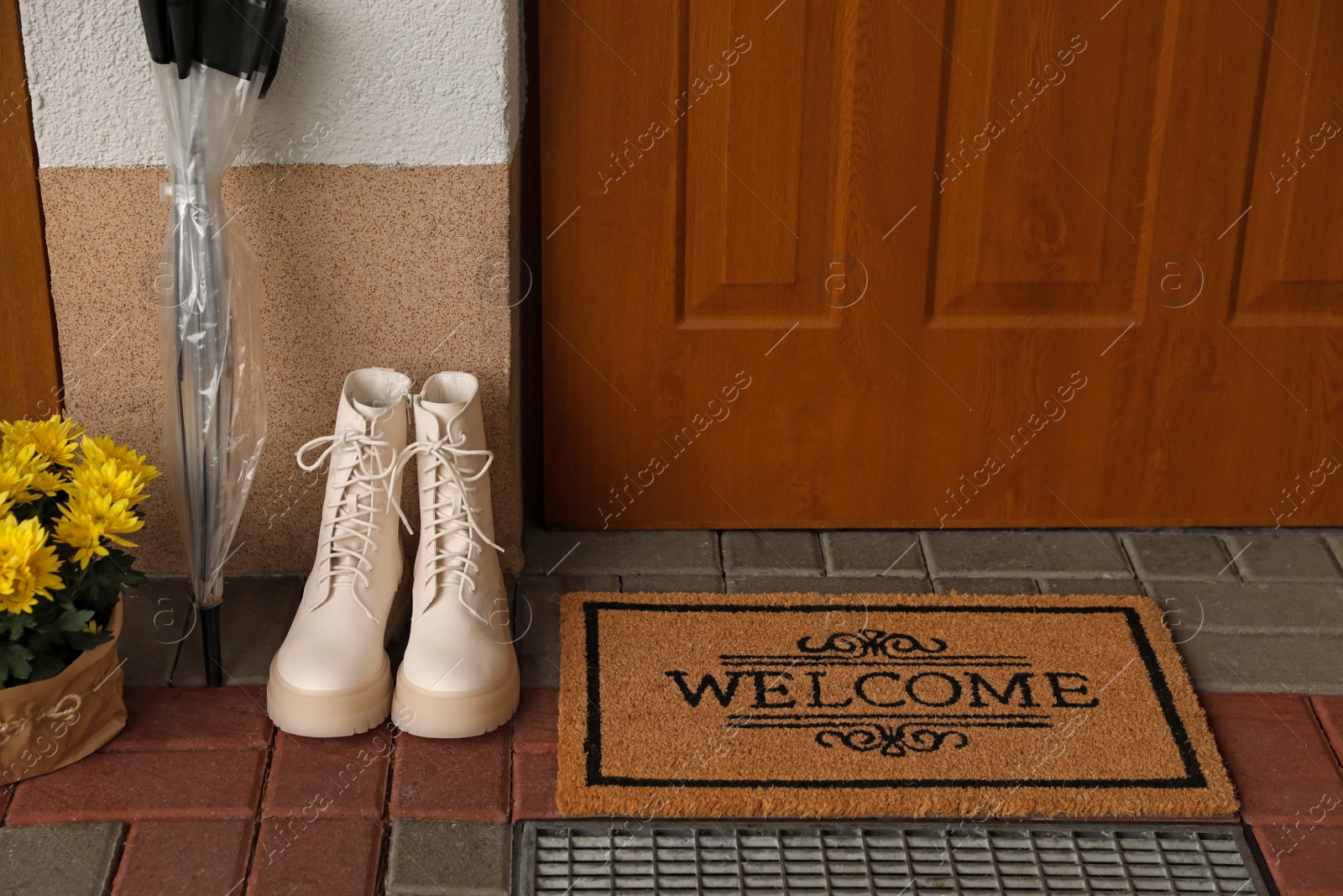 Photo of Door mat with word Welcome, boots, umbrella and beautiful flowers near entrance