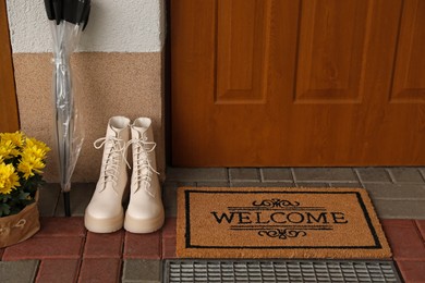 Photo of Door mat with word Welcome, boots, umbrella and beautiful flowers near entrance