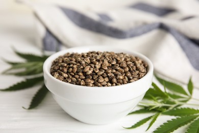 Bowl of hemp seeds and leaves on white table, closeup