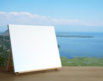 Image of Wooden easel with blank canvas on table and beautiful seascape. Space for text