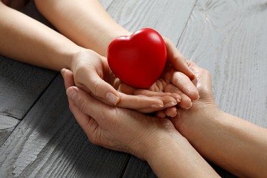 Photo of Young and elderly women holding red heart at grey wooden table, closeup