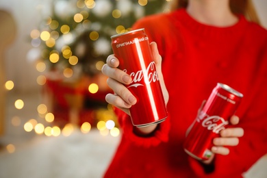 MYKOLAIV, UKRAINE - January 01, 2021: Woman with cans of Coca-Cola against blurred Christmas tree, closeup