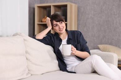 Beautiful young housewife with cup of drink on sofa at home