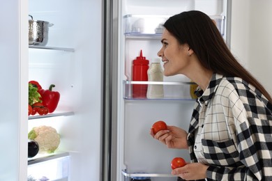 Photo of Young woman taking tomatoes out of refrigerator indoors