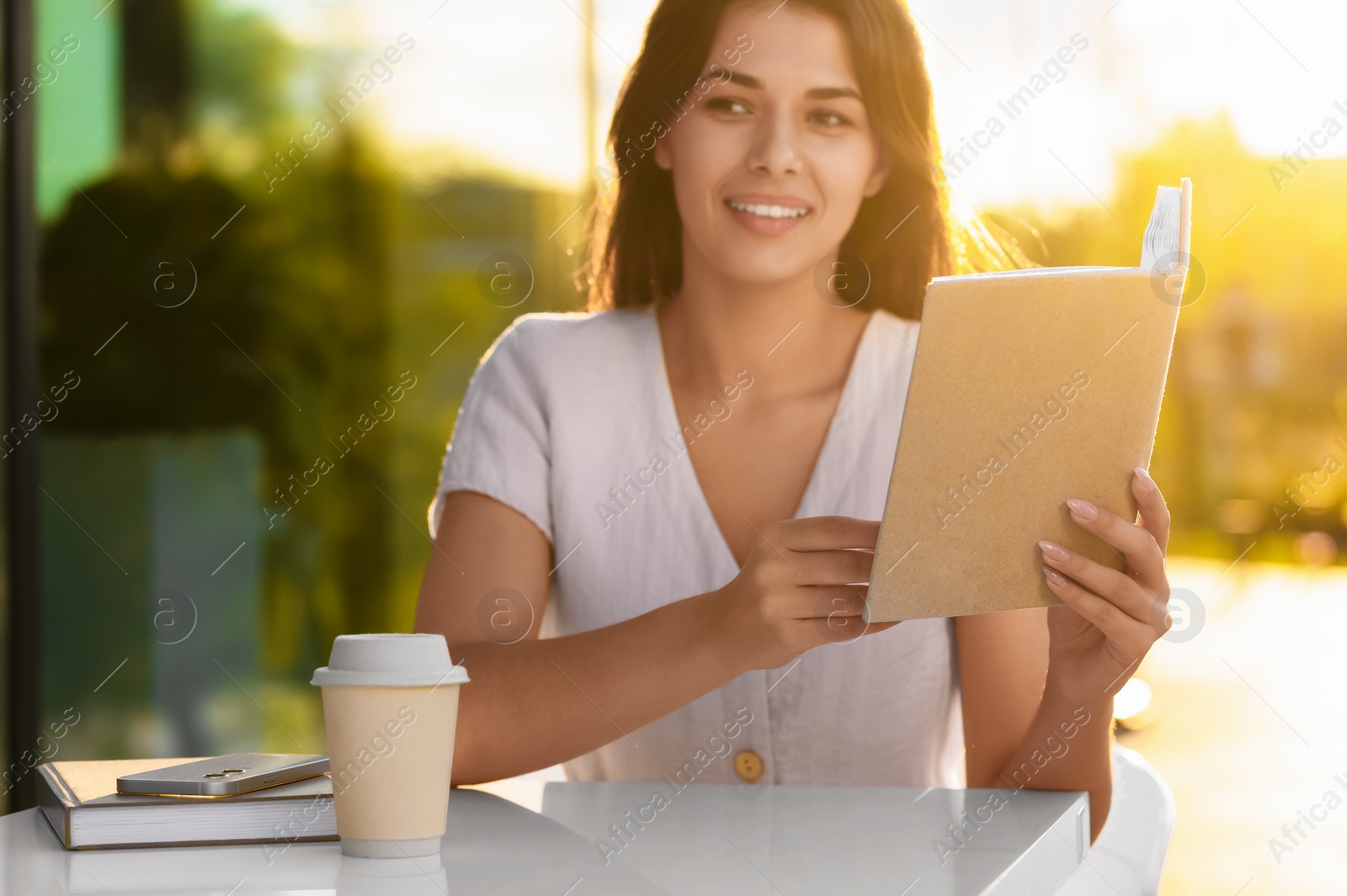 Photo of Young woman with coffee reading book in outdoor cafe on sunny day