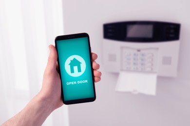 Man operating home alarm system via mobile phone against white wall with security control panel, closeup. Application interface on device screen