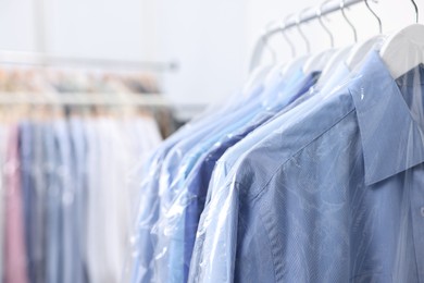 Photo of Dry-cleaning service. Many different clothes in plastic bags hanging on rack indoors, closeup